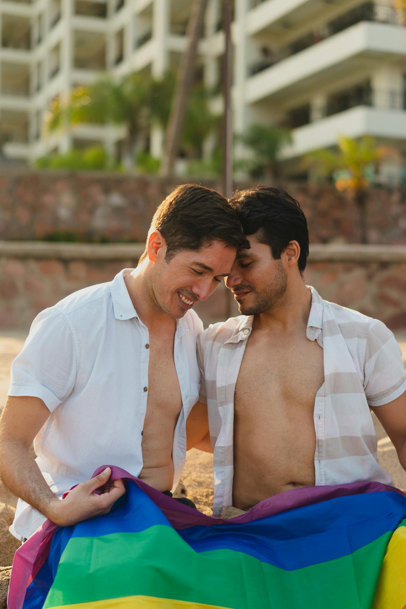 Couple enjoying a sunny day on the beach, draped in a rainbow flag.